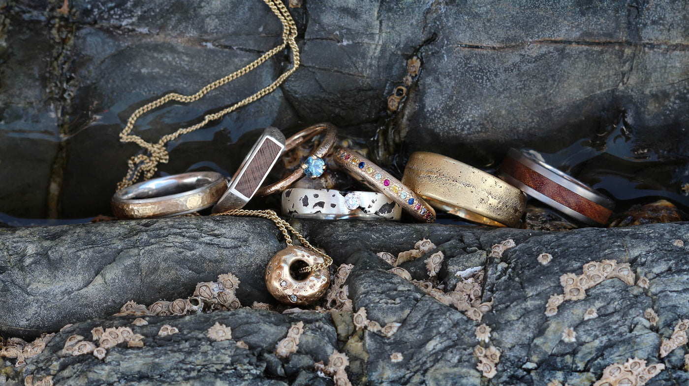 A selection of rustic, handmade rings and a pendant are displayed on a textured rock surface by the water's edge. The jewellery pieces, made from a mix of gold and silver, feature diverse finishes and designs—some with hammered textures, others adorned with colourful gemstones, wood inlays, or unique patterns. Small barnacles are visible on the rocks, adding an organic, seaside feel to the composition. A gold chain with a donut-shaped pendant lies in the foreground, enhancing the coastal aesthetic.